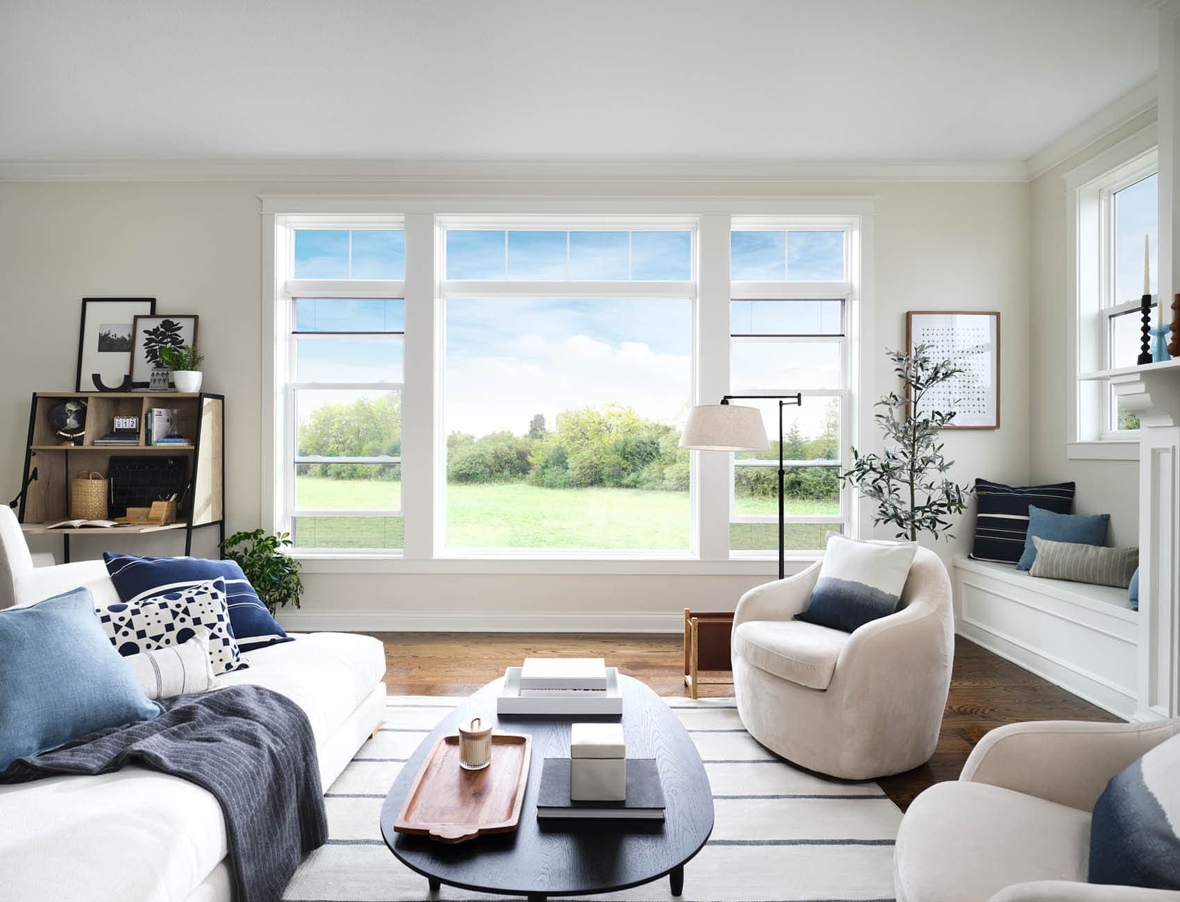 Bright, contemporary living room featuring a wide window wall with a large center fixed picture window flanked by two double-hung windows with top transom grilles. The space is styled with white walls, medium-tone wood floors, a cream sectional sofa accent with navy and light blue pillows, a round black coffee table, two plush cream swivel armchairs, and a built-in window bench seat along the side wall.
