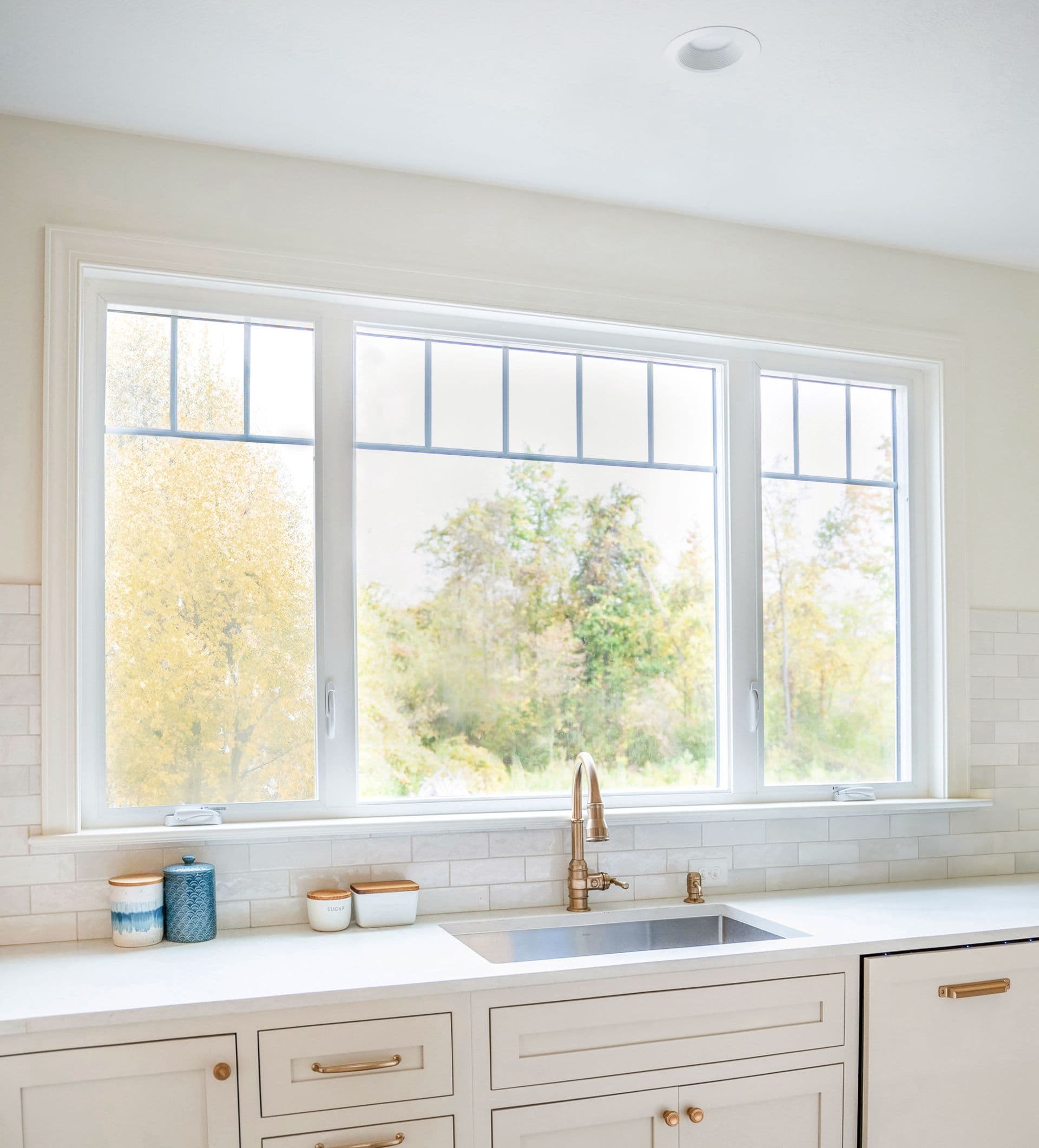 imgi_1_Aspect-Casement-Windows-Kitchen.jpg Interior view of a modern kitchen with white cabinetry, light countertops, white subway tile backsplash, and a brass faucet sitting in front of a wide three-lite white casement window system featuring top-grille accents and framing an outdoor view of autumn trees.