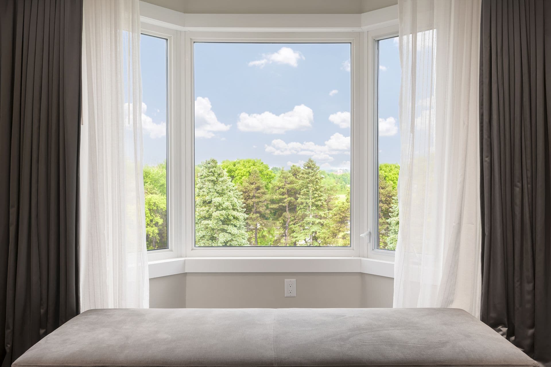 Bay window with drapes, curtains and view of trees under summer sky