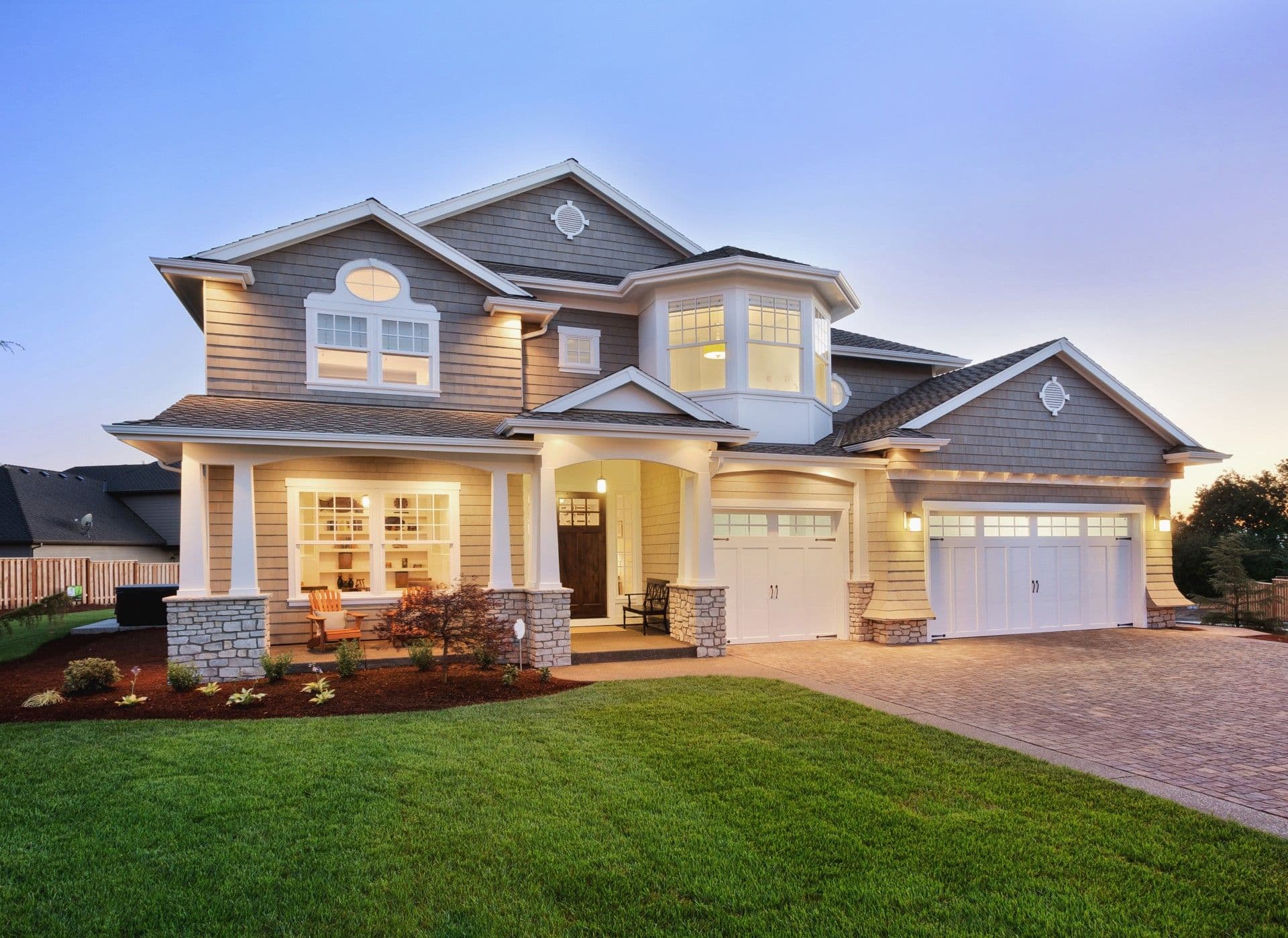 Two-story modern house at dusk with stone and siding exterior, illuminated windows, two white garage doors, front porch with columns, and manicured lawn