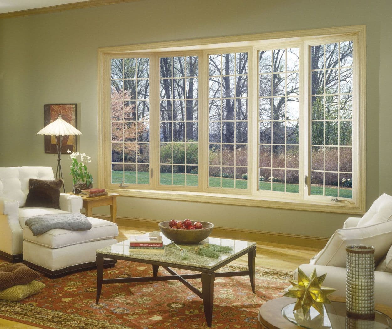 Interior view of a cozy living room featuring a large five-lite wooden bow window with decorative grilles, opening out onto a grassy yard with trees. The room is furnished with cream-colored armchairs, an ottoman, a coffee table on a patterned red oriental rug, and neutral wall paint.