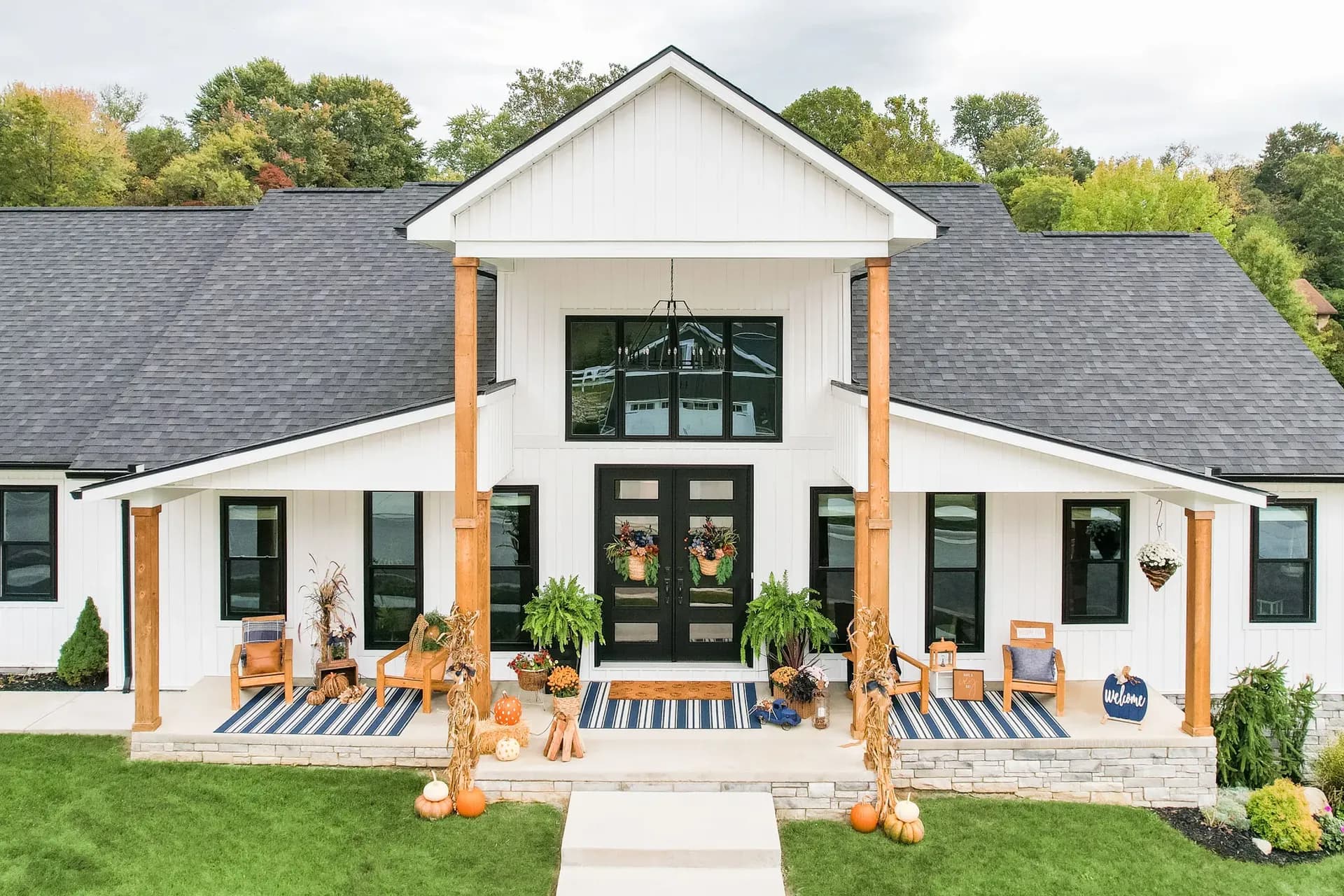 Modern farmhouse exterior with white siding, black double doors, wooden support columns, and fall decorations including pumpkins and corn stalks on the front porch