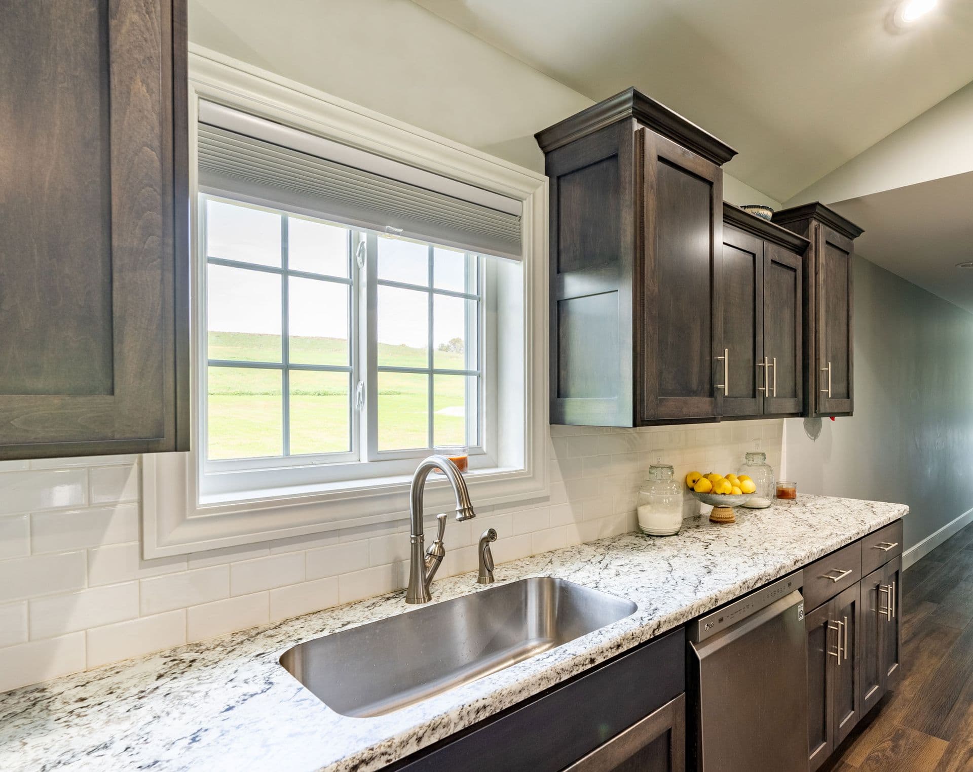 Interior view of a modern kitchen featuring dark-stained wood cabinetry, white granite countertops with dark veining, and a white subway tile backsplash. An undermount stainless steel sink sits beneath a white vinyl slider window with cottage-style grilles and a built-in cellular shade, offering a view of rolling green hills outside.