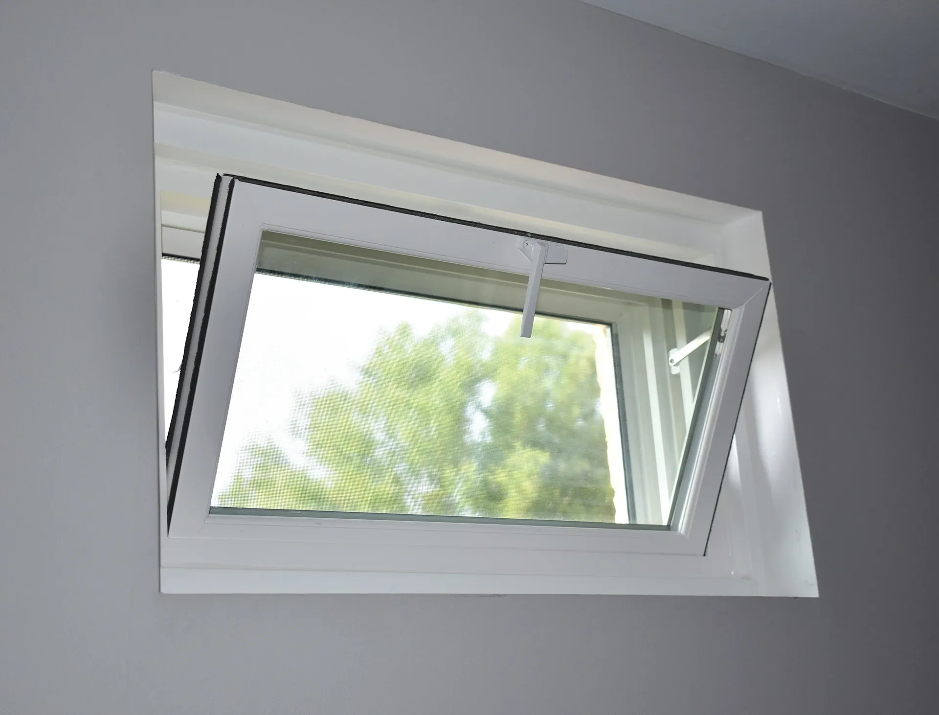 Interior view of a light grey wall featuring a modern white-framed hopper window, hinged at the bottom and tilted open inward at the top, equipped with a latch handle at the top sash and offering a view of green trees outside.