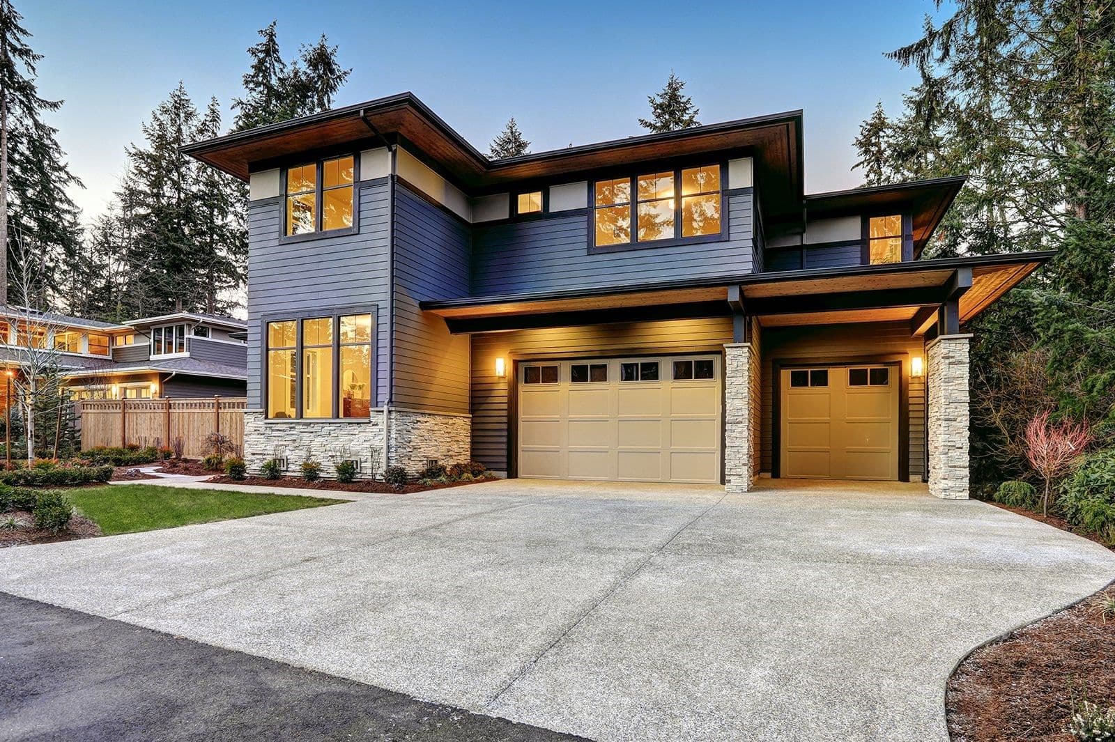 Modern two-story home with blue siding, stone accents, and two tan garage doors, surrounded by evergreen trees and lit windows at dusk