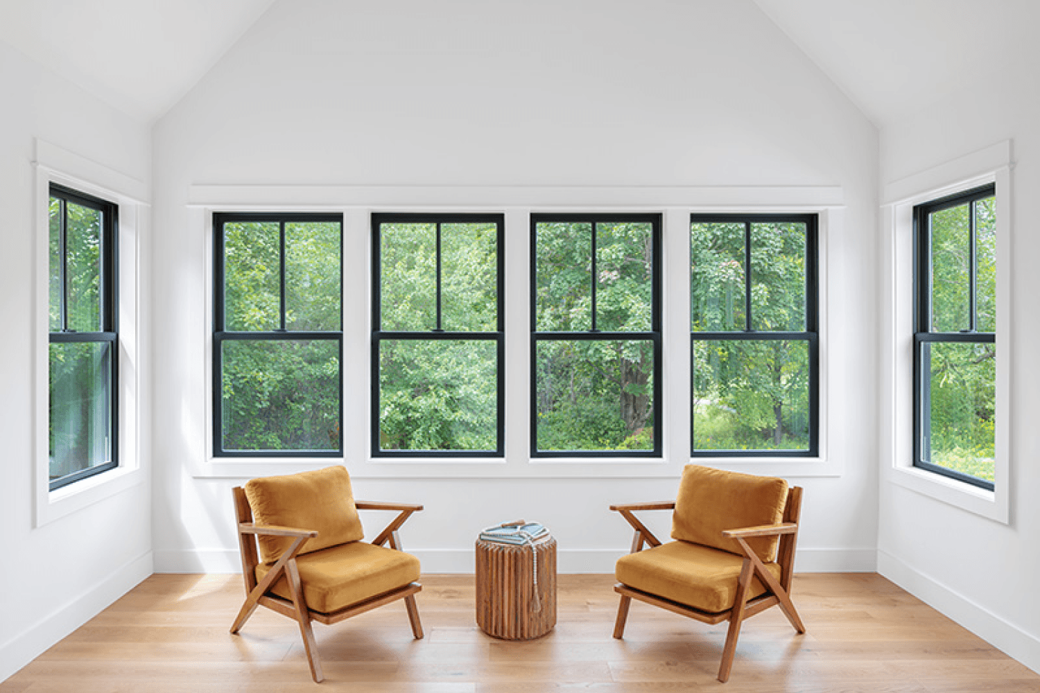 imgi_1_why-composite-windows_sunroom_3x2.jpg Interior view of a bright, minimalist sunroom with white vaulted ceilings, light wood flooring, and large black composite double-hung windows. Two mustard-yellow mid-century modern accent chairs face each other around a small carved wooden side table in front of a wide row of four windows overlooking vibrant green trees.